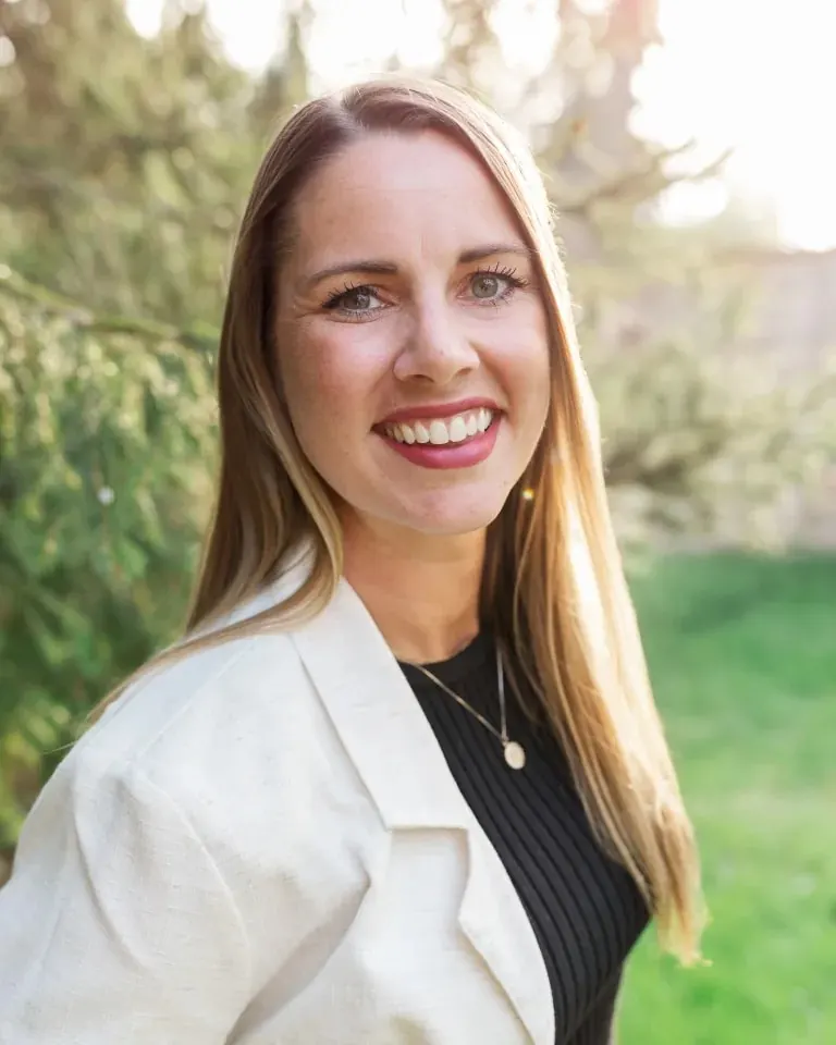 Bre Enbom outdoors in a white blazer, smiling, with evergreens behind her.