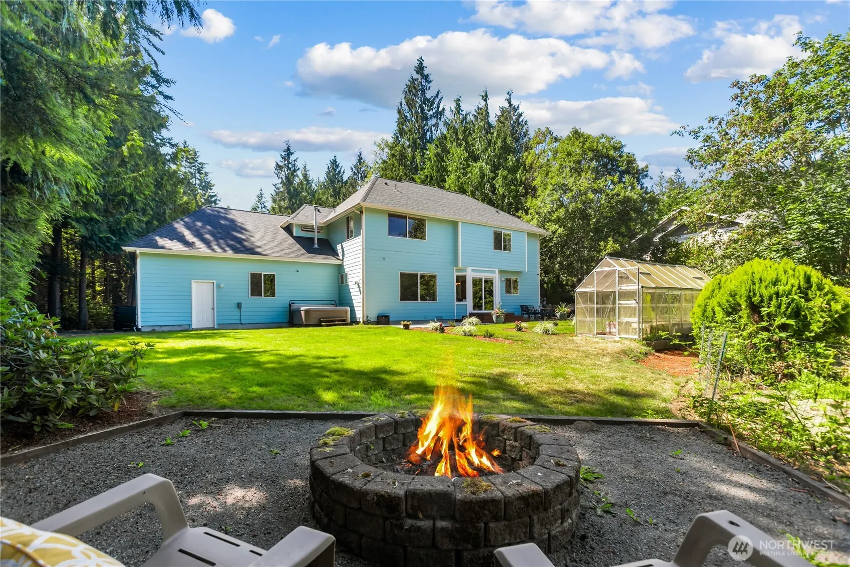 Stone fire pit with a fire lit, the lawn and greenhouse beyond, and the back of the house among tall trees.