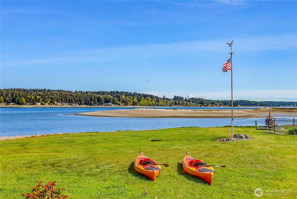 Waterfront lawn with two kayaks and a flag, a wooded shore and a snow-capped mountain beyond.